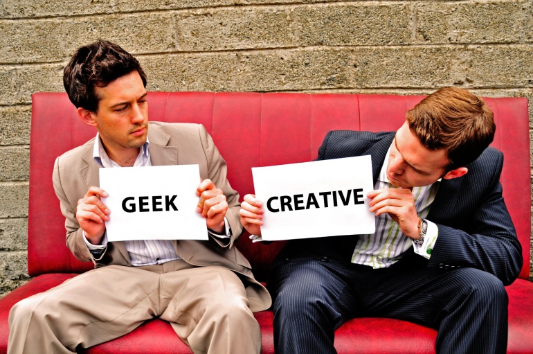 Patrick Cunningham holding a sign that says 'GEEK' sat next to Russell Isabelle holding a sign that says 'CREATIVE'