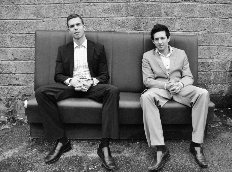 Russell Isabelle and Patrick Cunningham sat on a sofa wearing suits